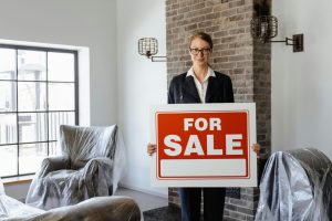 Professional real estate agent holding a 'For Sale' sign in a furnished indoor setting.